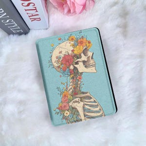 Puede incluir: Una funda rectangular para lector electrónico con fondo azul claro que presenta una ilustración detallada de un cráneo humano y la parte superior de la caja torácica, adornada con flores amarillas, naranjas, rojas y rosas.