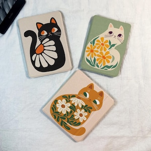 Puede incluir: Tres fundas rectangulares con ilustraciones de gatos. Una funda presenta un gato negro con un diseño de margarita, otra tiene un gato blanco con flores amarillas y la tercera un gato naranja con flores blancas.