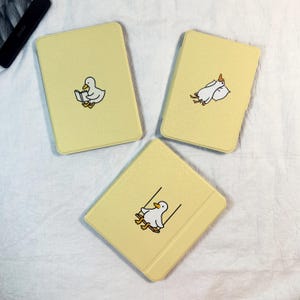 Puede incluir: Tres fundas amarillas para Kindle con ilustraciones de patos. Una muestra un pato leyendo un libro, otra un pato descansando sobre una almohada y la tercera un pato en un columpio. Las fundas son rectangulares con esquinas redondeadas.