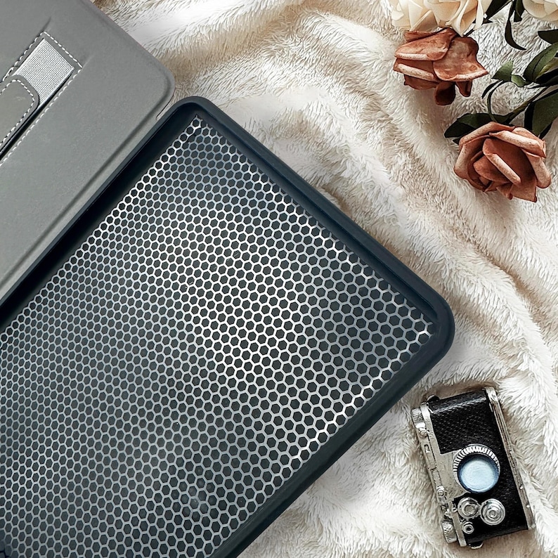 May include: A dark gray tablet case with a honeycomb pattern, a gray tablet cover, and a vintage camera are displayed on a soft, white, textured surface. Dried roses are in the background.