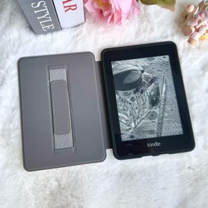 May include: A black Kindle e-reader with a gray cover, open to display an image of an inkwell and pen. The cover has a built-in stand. The word "Kindle" is printed on the bottom of the device.