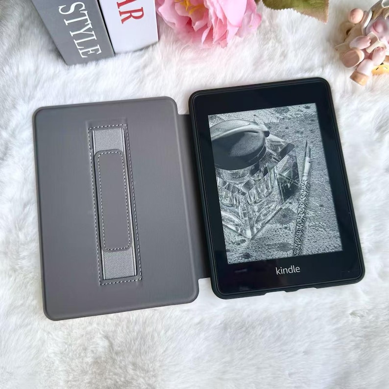 May include: A black Kindle e-reader with a gray cover, open to display an image of an inkwell and pen. The cover has a built-in stand. The word "Kindle" is printed on the bottom of the device.