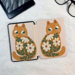 Pode incluir: Uma capa para leitor eletr&oacute;nico com fundo rosa claro e duas ilustra&ccedil;&otilde;es id&ecirc;nticas de um gato laranja. O gato tem olhos verdes e est&aacute; decorado com flores de margarida brancas e folhas verde-escuras no corpo e na cauda.