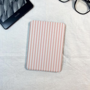 May include: A pink and white striped Kindle cover lies on a white surface, alongside a Kindle device and black-framed glasses. The cover features a vertical stripe pattern.