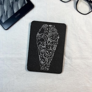 Puede incluir: Posavasos rectangular negro con una ilustración blanca de elementos temáticos de Halloween, incluyendo una calavera, una calabaza, un cuchillo y un reloj de arena. El posavasos está sobre una superficie blanca, con un Kindle y gafas en el fondo.