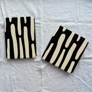 May include: Two rectangular coasters featuring a high-contrast pattern of vertical, rounded off-white shapes set against a solid black background. The coasters have smooth, rounded corners and a flat surface, displaying a consistent graphic design.