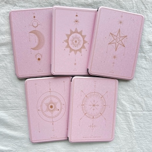 Pode incluir: Cinco baralhos de cartas de tarot rosa com desenhos celestiais dourados, incluindo uma lua crescente, um sol e uma estrela. As cartas estão dispostas em forma de leque, com os títulos "MOON", "SUN", "STAR", "THE WORLD" e "WHEEL OF FORTUNE".