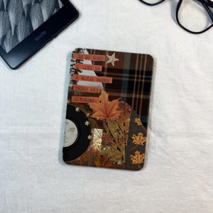 May include: A rectangular e-reader skin featuring a collage design with a brown plaid pattern, a black vinyl record, and autumn maple leaves. The text reads: I'M SO GLAD I LIVE IN A WORLD WHERE THERE ARE OCTOBERS. The skin has a matte finish and a star motif.