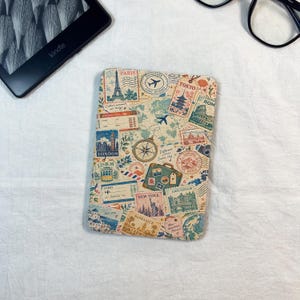 May include: A rectangular e-reader cover featuring a vintage travel-themed collage. The design includes illustrations of the Eiffel Tower, the Statue of Liberty, and various city names like Paris, Tokyo, London, Lisbon, New York, and Amsterdam.