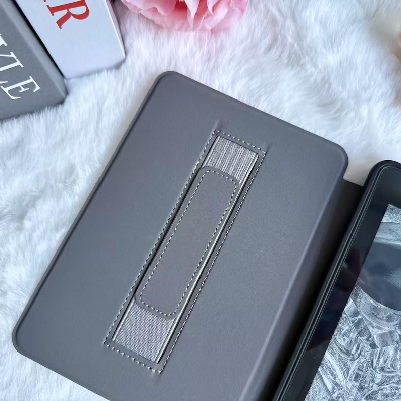 May include: Gray tablet case with a silver elastic hand strap. The case is open, revealing a portion of the tablet screen. The case is on a white, fluffy surface, with a pink flower and a book visible in the background.