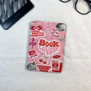 Op de afbeelding: Een rechthoekige e-readerhoes met een roze rasterachtergrond, versierd met diverse stickers waaronder kersen, een discobal, een speelkaart en teksten zoals "Book Lover", "One More Chapter", "Hot Girls Read" en "Emotional Support Kindle".