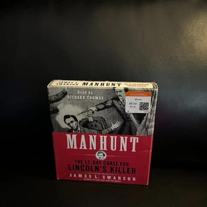 May include: A red and white book titled "MANHUNT" by James L. Swanson, detailing the 12-day chase for Lincoln's killer. The cover features illustrations and the text "Read by Richard Thomas."
