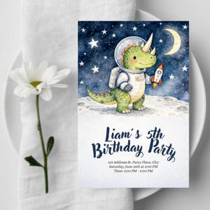 Puede incluir: Una invitación de cumpleaños en acuarela con un dinosaurio astronauta verde sosteniendo un cohete contra un cielo estrellado. La invitación dice "5º Cumpleaños de Liam" con los detalles del evento a continuación.