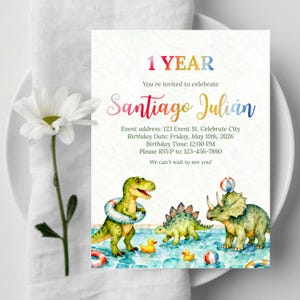 Peut inclure: Une invitation d'anniversaire avec une illustration à l'aquarelle de dinosaures dans une piscine. L'invitation indique "1 YEAR" et invite les invités à célébrer l'anniversaire de Santiago Julián le 10 mai 2026 à 12h00. Les informations RSVP sont incluses.