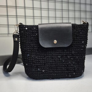 May include: Black crochet shoulder bag with a flap closure. The bag features a black leather flap secured with a gold-tone button. The bag has a black strap and is embellished with sequins.
