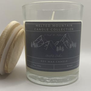 May include: A glass jar candle with a black and white label that reads "Melted Mountain Candle Collection - Find It & Explore -  Adirondack Cabin - Soy Wax Candle - 7 oz / 200g - 55 Hours".