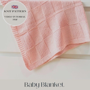 May include: A pink knitted baby blanket with a square pattern. The blanket is folded and draped over a white surface. The text "KNIT PATTERN VIDEO TUTORIAL PDF" is visible in a circle at the top of the image.