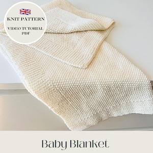 Op de afbeelding: Een crèmegekleurde gebreide babydeken. De deken is opgevouwen en toont een gestructureerd steekpatroon. De afbeelding bevat een cirkelvormige afbeelding met de tekst "KNIT PATTERN VIDEO TUTORIAL PDF", de Britse vlag en de woorden "Baby Blanket".