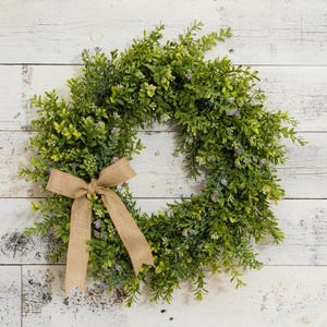 May include: A green and purple artificial wreath with a brown burlap bow. The wreath is made of faux greenery and has small purple flowers.