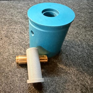 May include: A teal blue cylindrical object with a hole at the top, next to a smaller, light gray cylindrical object with a gold-colored rod attached. The objects are on a gray surface.