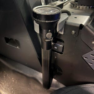 May include: A black flashlight with a ribbed handle is mounted in a vehicle. The flashlight has a clear lens and a gray button. The mount is gray and attached to a black console. The vehicle's interior includes air conditioning controls.
