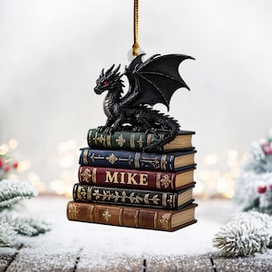 May include: A black dragon ornament with red eyes and large wings sits atop a stack of antique-style books. The books are various colors, including green, blue, and brown, with the name "MIKE" on one. A gold string is attached for hanging.