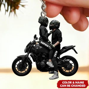 May include: A black motorcycle keychain featuring two figures wearing helmets and black outfits, seated on the bike. The text "SOPHIA" and "ALEX" are visible on the figures' clothing. The keychain has a red banner that reads "COLOR & NAME CAN BE CHANGED."
