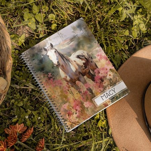 Peut inclure: Un carnet à spirales avec une peinture à l'aquarelle d'un cheval sur la couverture. Le carnet est posé sur un lit d'herbe verte avec un chapeau brun et un panier de pommes de pin à proximité. Le carnet porte le nom "MACY" imprimé sur la couverture.