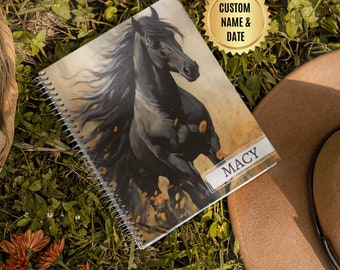 May include: A black horse with a flowing mane runs through a field of yellow flowers. The image is on a spiral bound notebook with the name "MACY" printed on the cover.  The notebook is on a bed of green grass.