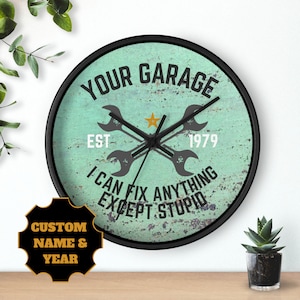 May include: A teal blue round clock with a black frame. The clock face has a distressed metal look with the text "YOUR GARAGE EST 1979 I CAN FIX ANYTHING EXCEPT STUPID". There are two crossed wrenches on the clock face.