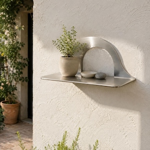 Pode incluir: Uma prateleira de parede em metal escovado com uma placa traseira em arco semicircular. A superfície horizontal plana contém um pequeno vaso de cerâmica bege com uma planta verde, uma pequena tigela branca rasa e uma pedra cinzenta lisa.
