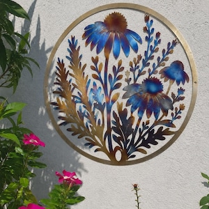 May include: A circular metal wall art piece featuring a floral design with two large daisy-like flowers and surrounding foliage. The metal has a heat-treated finish, displaying vibrant gradients of blue, purple, and golden-brown tones throughout.