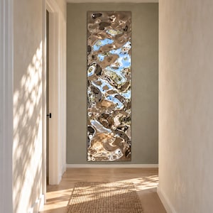Puede incluir: Un espejo de pared alto y rectangular con una superficie metálica ondulada y muy pulida. El acabado reflectante crea una apariencia líquida y distorsionada, capturando el cielo azul y los reflejos de luz en su textura plateada.