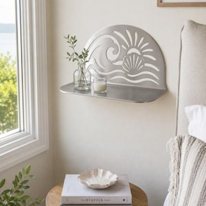 Op de afbeelding: Een zilverkleurige metalen wandplank met een halfrond achterpaneel met een uitgesneden ontwerp van een gestileerde oceaangolf, een zon en een schelp. Het plankoppervlak is plat en rechthoekig en bevat een glazen vaas met groen en een kaars.