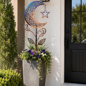May include: A metal garden stake featuring a crescent moon with a filigree pattern and a hanging star, finished in iridescent blue and copper tones. The stake has a central stem with decorative leaves and is positioned inside a tall gray planter.
