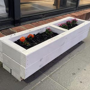 May include: A white wooden rectangular planter with two sections, filled with dark soil and colourful orange and pink flowers. The planter sits on grey paving slabs next to a brick wall.