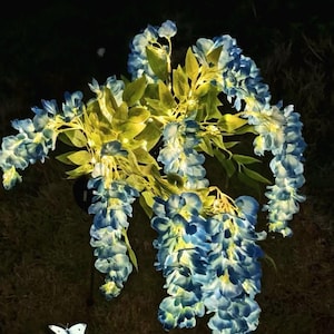 Puede incluir: Un racimo de flores de glicina artificiales en tonos azules y blancos, con hojas verdes. Una mariposa blanca está en la esquina inferior izquierda. La glicina está iluminada sobre un fondo oscuro.