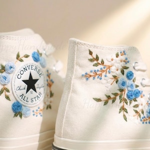 Puede incluir: Zapatillas altas color crema con intrincado bordado floral. El diseño presenta rosas azules, flores blancas y hojas verdes. El logotipo de Converse All Star es visible.