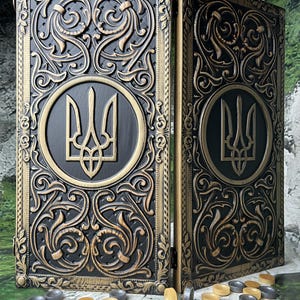 May include: A black and gold backgammon set with ornate carved detailing. The set features a Ukrainian trident emblem. The game pieces are gray and tan, with two blue dice. The set is displayed against a natural background.