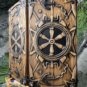 Könnte beinhalten: Ein klappbares Backgammon-Spielbrett aus Holz mit detaillierten Schnitzereien eines Schiffssteuerrads, Ankern und Ketten. Das Brett hat ein zweifarbiges Design mit natürlicher Holzmaserung und schwarzen Akzenten. Runde Spielsteine liegen davor.