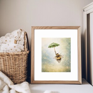 K&ouml;nnte beinhalten: Gerahmter Kunstdruck mit einer Illustration einer Hummel, die einen gr&uuml;nen Regenschirm mit wei&szlig;en Punkten h&auml;lt. Die Hummel fliegt vor einem vertr&auml;umten Hintergrund. Das Bild ist in einem hellen Holzrahmen gefasst.
