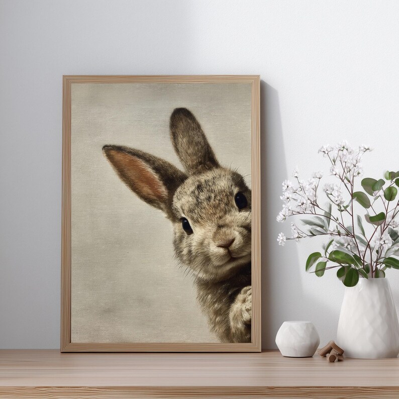May include: A framed print featuring a detailed illustration of a rabbit peeking from the side. The rabbit has brown and gray fur, with long ears and dark eyes. The artwork is set against a neutral background, and the frame is made of light-colored wood.