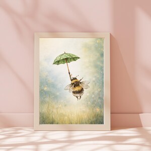 K&ouml;nnte beinhalten: Gerahmter Kunstdruck mit einer Illustration einer Hummel, die einen gr&uuml;nen Regenschirm mit wei&szlig;en Punkten h&auml;lt und &uuml;ber einer Blumenwiese schwebt. Das Kunstwerk hat einen weichen Aquarellstil mit hellem, luftigem Hintergrund und beigem Rahmen.
