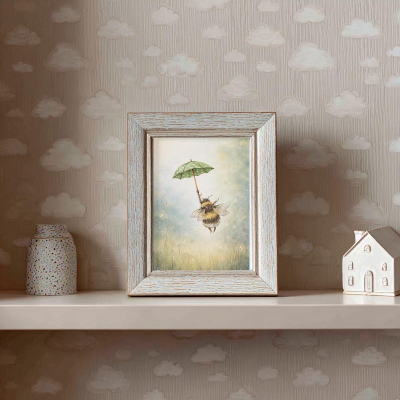 K&ouml;nnte beinhalten: Ein gerahmter Kunstdruck mit einer Illustration einer Hummel, die einen gr&uuml;nen Regenschirm h&auml;lt, vor einem weichen, vertr&auml;umten Hintergrund. Der Rahmen hat eine verwitterte, hellblaue Oberfl&auml;che. Der Druck wird auf einem Regal mit einer dekorativen Vase und einem kleinen wei&szlig;en Haus ausgestellt.