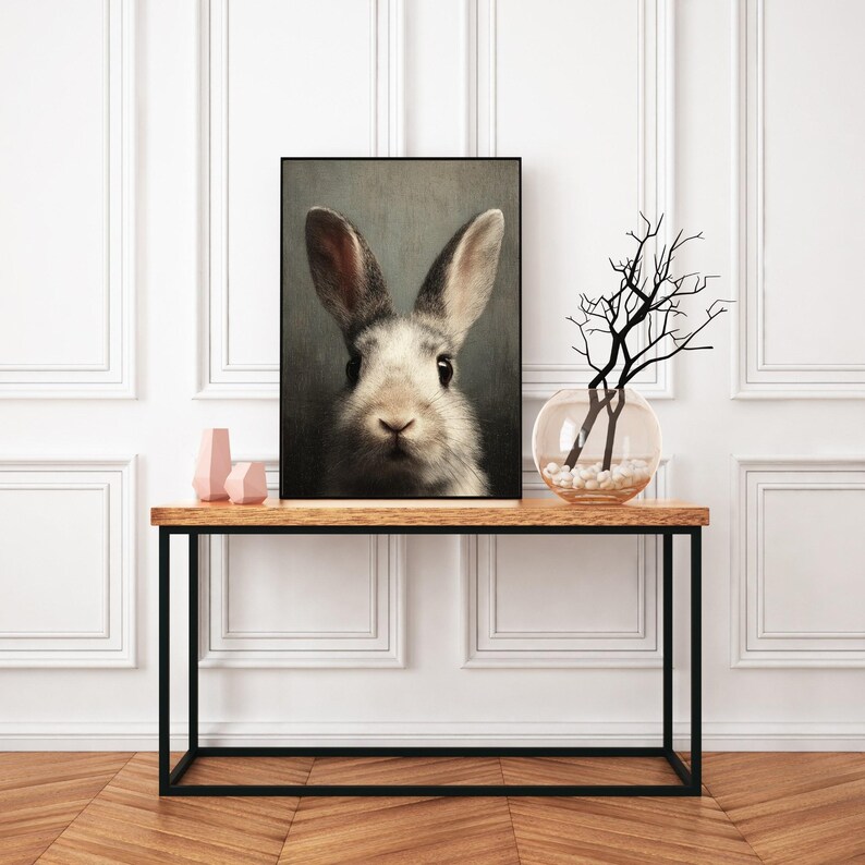 Op de afbeelding: Een ingelijst portret van een konijn met een zwart frame. Het konijn heeft witte en grijze vacht, grote oren en donkere ogen. Het kunstwerk wordt tentoongesteld op een houten tafel met een zwart metalen frame, met decoratieve items.