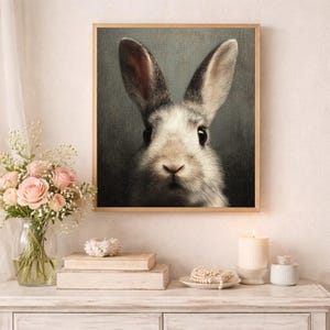 Op de afbeelding: Een ingelijst portret van een konijn met zachte, witte en grijze vacht. De grote, donkere ogen en lange oren van het konijn zijn het middelpunt van het kunstwerk. De houten lijst voegt een rustieke toets toe aan het stuk, geschikt voor interieurdecoratie.