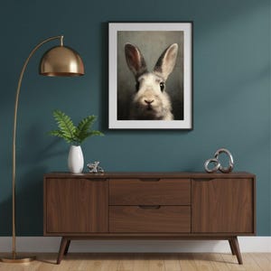 Op de afbeelding: Een ingelijste print van een konijnengezicht hangt boven een houten kast. De kast heeft twee lades en twee deuren. Een gouden vloerlamp en decoratieve items, waaronder een vaas met groen, zijn ook zichtbaar in de kamer. De muur is tealblauw geschilderd.