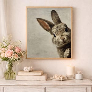 May include: A framed print featuring a detailed illustration of a rabbit peeking from the side. The rabbit has brown and gray fur, with long ears and a curious expression. The artwork is set in a light-colored wooden frame, suitable for home decor.