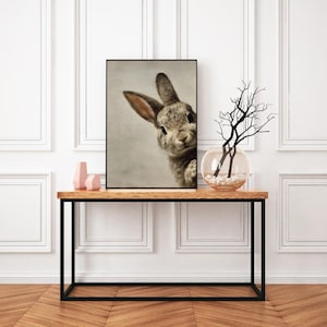 May include: A framed print featuring a close-up of a curious rabbit. The rabbit has gray and brown fur, with long ears and a sweet expression. The print is displayed on a wooden table with a black metal frame, alongside decorative items.
