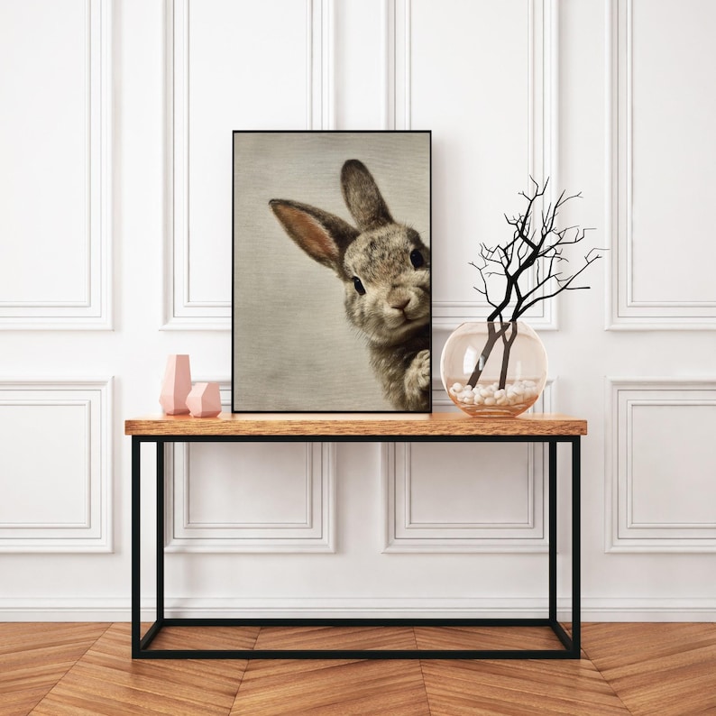 May include: A framed print featuring a close-up of a curious rabbit. The rabbit has gray and brown fur, with long ears and a sweet expression. The print is displayed on a wooden table with a black metal frame, alongside decorative items.